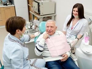 An older, smiling man pointing at his mouth 